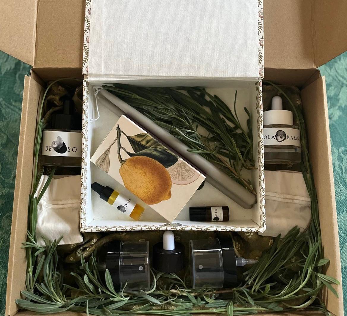 Caja de cartón abierta con ramitas de romero, dos velas, frascos de perfume y un estuche decorado con motivos botánicos, sobre una tela verde oliva